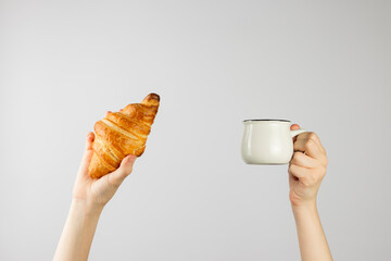 Girl hand holding white ceramic coffee cup and croissant on light background.