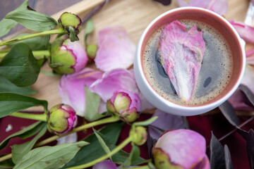 A white Cup of black coffee stands on a wooden background next to pink peonies and other garden flowers and leaves, The concept of village life, Cottagecore, ruralcore, cluttercore. High quality photo