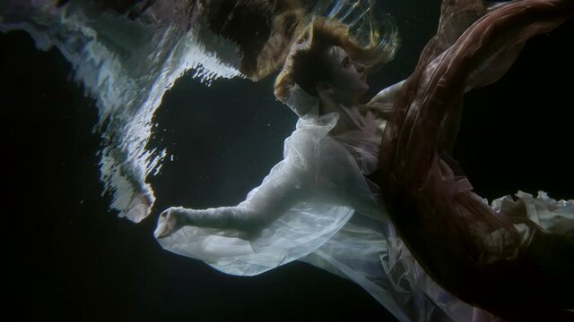 a woman with red hair swims in the dark under water with a translucent cloth like a mermaid in a fairy tale