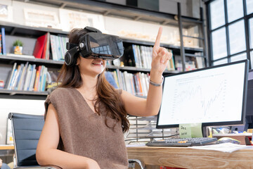 Happy Asian office lady wearing a virtual reality headset and make a gesture touching something in the air while sitting at desk with computer showing a statistic graphs on screen in a blurred office.