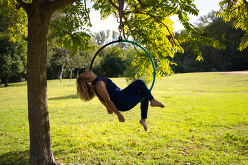 Blonde woman and young gymnast acrobat athlete performing aerial exercise on air ring outdoors in park. Lithe woman in blue costume performs poses of circus performers dancing with hips