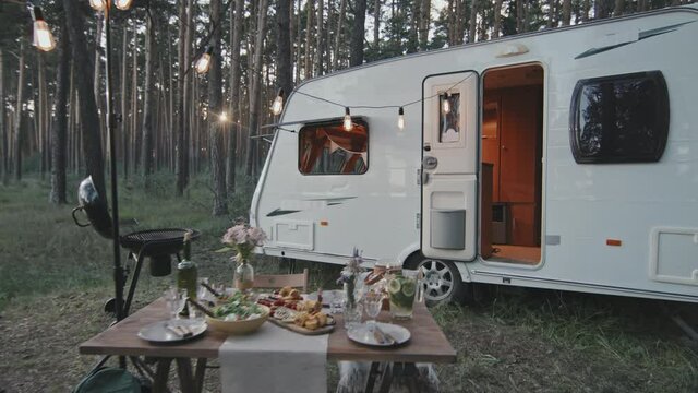 Dolly-out tracking of white campervan parked in forest and table laden with delicious food