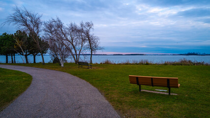 The curving footpath and an empty bench in the park with bare trees. Moody cloudscape at dusk. Twilight seascape over the green at Nut island Pier Park in Quincy, Massachusetts.