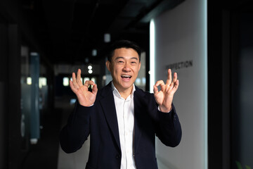 Successful businessman rejoices looking at camera shows thumbs up and signs ok, asian boss happy and laughs