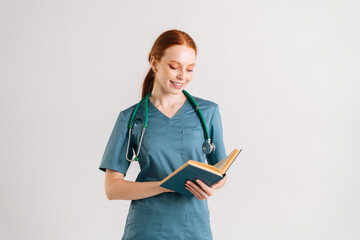 Portrait of smiling young woman intern in green uniform with stethoscope reading medical book...