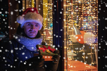 Obraz na płótnie Canvas Man in Santas hat with gifts box near illuminated cafe window.