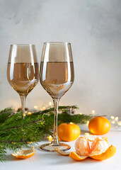 Two transparent glasses of champagne on a light table. Tangerines, fir branches and garlands in the background. Festive atmosphere