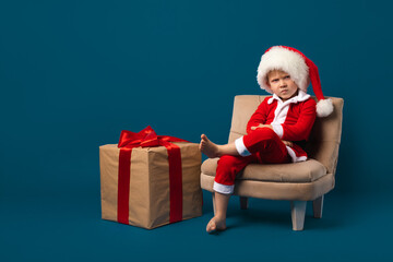 Child boy in bad santa costume with New Year hat, christmas concept. Big gift box with red ribbon....