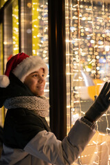 Fototapeta na wymiar Boy in Santas hat looking and dreaming in illuminated shop window. Xmas presents holidays, or shopping on New Year or Christmas concept