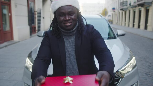 Young black man with santa hat giving red wrapped christmas gift in front of his car. High quality 4k footage