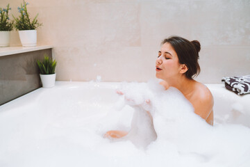 Young woman lies in white bath of foam with her eyes closed. Spa procedures, cleansing, relaxation, self-care.