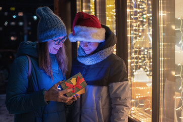 Pretty woman and her son in santas hat opening gift box on illuminated shop window background. New Year or Christmas concept