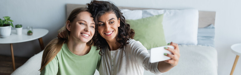 curly woman taking selfie with happy girlfriend in bedroom, banner