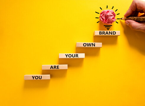 You are your own brand symbol. Wooden blocks with words You are your own brand. Beautiful yellow background, copy space. Businessman hand, light bulb. Business, you are your own brand concept.