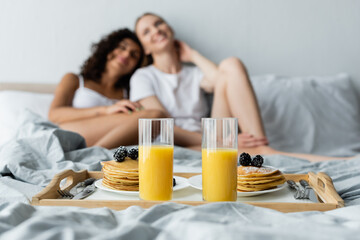 Obraz na płótnie Canvas glasses with orange juice and pancakes on tray near blurred lesbian couple