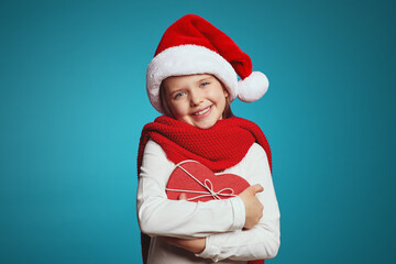 Portrait of cheerful caucasian kid dressed in christmas hat, red scarf embracing present box in shape of heart, isolated over blue background 