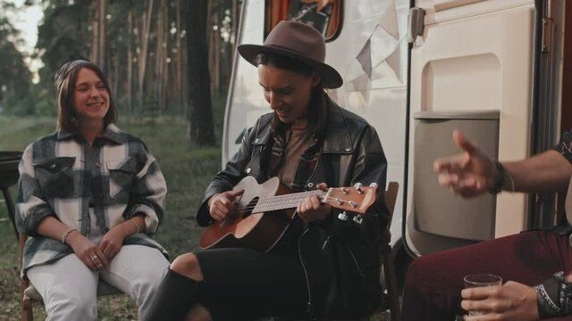 Handheld slowmo of young woman sitting before campervan parked in forest and playing ukulele while her cheerful friends singing song and enjoying music