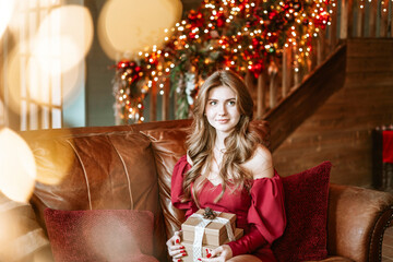 beautiful young woman in red dress sits on couch near Christmas tree in living room decorated for celebration of Christmas and New Year and opens boxes with wrapped Christmas presents