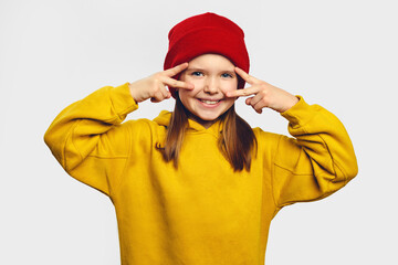Happy optimistic teenage girl makes peace victory sign over eyes, disco gesture, has fun, wears yellow hoodie and red hat over white background 
