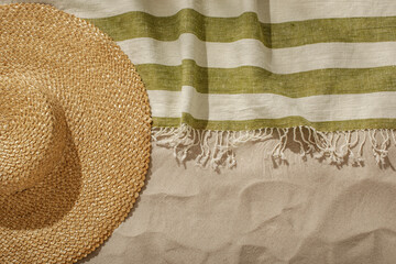Striped linen beach towel with fringes and straw hat on sandy beach with shadows from palm tree. Relaxation and tropical summer holidays concept