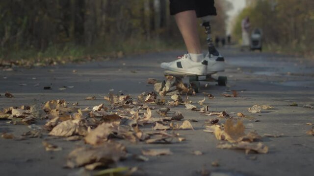 A young man with a metal bionic prosthetic leg is riding a skateboard in the autumn forest. An artificial leg pushes off the asphalt on a skateboard. A full life of a disabled person in the park.