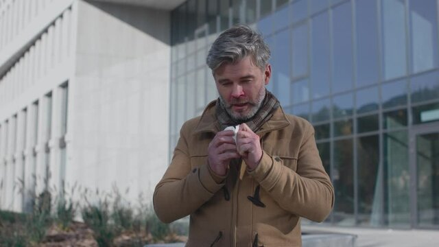 Sick senior man sneezing on city street background. Ill male with cold symptoms sneeze outdoors while using napkin. Portrait of the male suffering from allergy and feeling bad