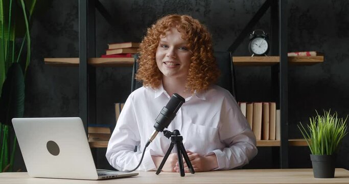 Smiling influencer red-haired woman recording video porcast with professional microfone. Digital blogger vlogger streaming talk show in home office studio, sitting at desk.