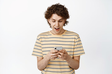 Excited curly guy using mobile phone screen, texting message on smartphone, standing over white background