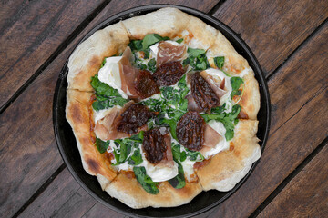 Top view of an arugula pizza with burrata cheese, sun dried tomatoes and cured raw ham, served in the pizza pan on the wooden table. 