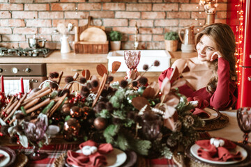 beautiful young woman in red dress with glass of wine stands near Christmas tree in living room decorated for celebration of Christmas and New Year in stylish interior, table set for family dinner