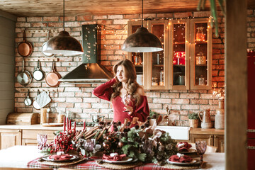 beautiful young woman in red dress with glass of wine stands near Christmas tree in living room decorated for celebration of Christmas and New Year in stylish interior, table set for family dinner