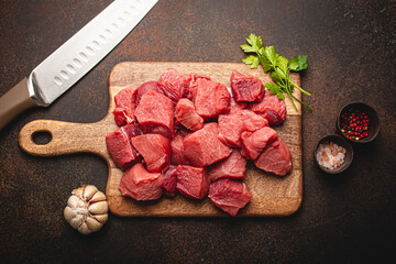 Raw beef meat chopped in cubes with bunch of fresh parsley, garlic, salt and pepper on wooden cutting board with knife for cooking stew or meat dish on brown dark stone background top view flat lay