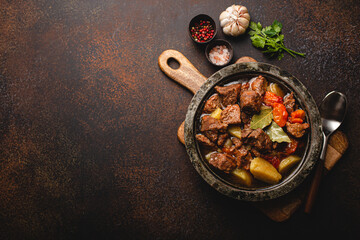 Delicious beef meat stew dish with potatoes, carrot and gravy in rustic metal bowl with spoon, bunch of fresh parsley, garlic cloves, spices on brown concrete background top view space for text - obrazy, fototapety, plakaty