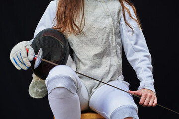 Close up of a fencer dressed in white fencing, holding his mask and a sword on black background.