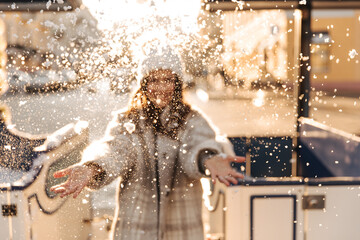 Photo of fair-skinned girl, covered with flakes of snow, standing by carriage on street. Brunette in hat and coat is having fun before coming winter holidays.