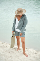 Young girl in natural linen clothes and a straw hat, barefoot on a white sand beach, holding a linen bag. Back view.