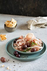 Slices of thinly sliced ham (prosciutto, Parma ham) with a bowl of olives on a light gray background. An appetizing Mediterranean snack, antipasti. 