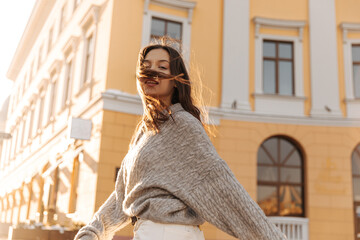 Beautiful fair-skinned young woman poses vigorously against backdrop city building on warm day. Her dark hair flying in wind, she dressed autumn grey clothes. Leisure concept youth, active lifestyle