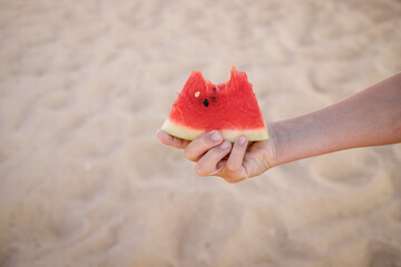 A cut slice of watermelon in a child's hand against the backdrop of a sandy beach.