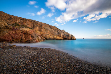 Photograph taken with long exposure on a beach with crystal blue waters in Cartagena, in the region of Murcia, Spain on a sunny day, with boulders on the shore and a boat on the horizon.