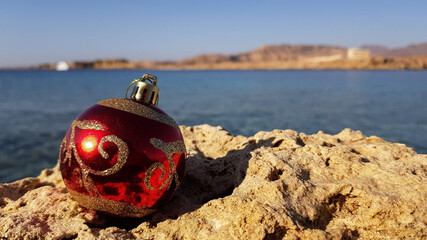Christmas tree decoration in the form of a red ball on the background of the sea and the beach. Christmas in warm countries
