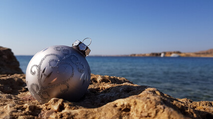 Christmas tree toy in the form of a ball of silver color on the background of the beach and the sea. Christmas on the beach in a warm country