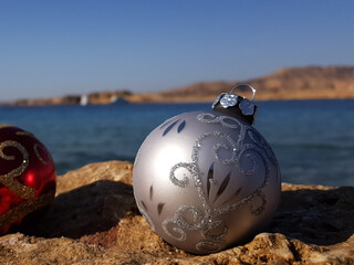 Christmas tree toy in the form of a ball of silver color on the background of the beach and the sea. Christmas on the beach in a warm country