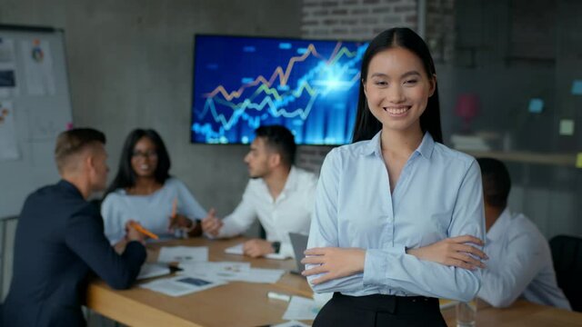 Young positive asian business lady smiling to camera with folded arms, enjoying her work with diverse colleagues team