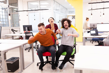 Fototapeta na wymiar Colleagues having fun on work at the office. Girls pushing boys on chairs with wheels. Multiethnic group of successful start up company.
