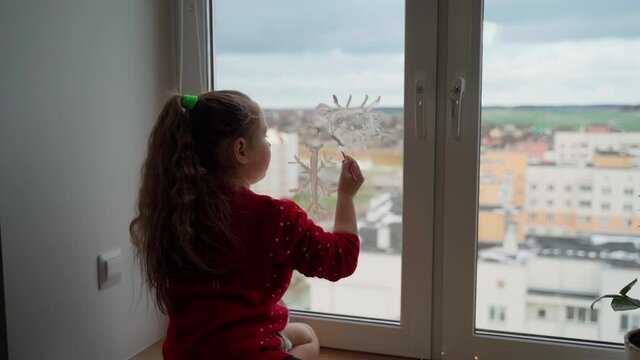 Child draw santa's gnome on window with white paints close-up. Caucasian female person decorate living room for christmas holidays and having fun indoors. Cute girl prepare to new year at home