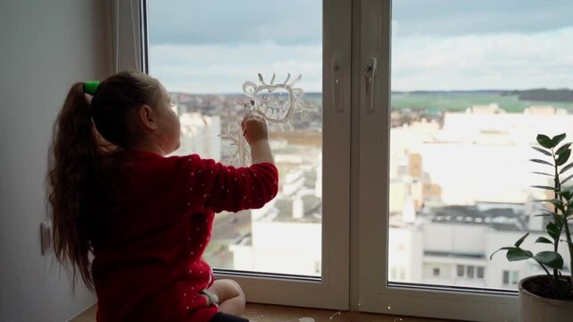 Child draw santa's gnome on window with white paints close-up. Caucasian female person decorate living room for christmas holidays and having fun indoors. Cute girl prepare to new year at home