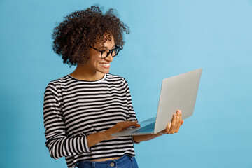 Joyful multiethnic lady in glasses holding silver laptop and smiling. Isolated on blue background