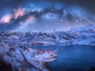 Milky Way above sea coast, snow covered mountains, village with red rorbu in winter at night in...
