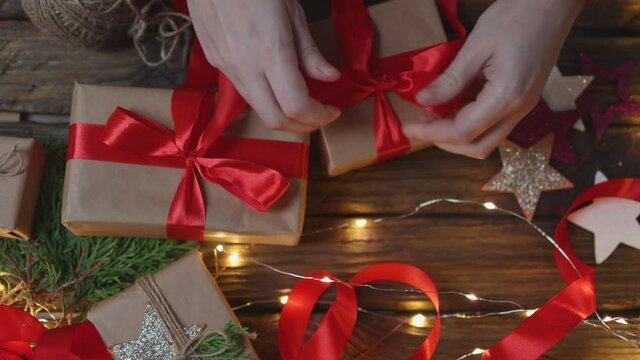 female woman hands wrapping Christmas presents gift box with brown craft paper, tying red ribbon bow on wooden desk with new year ornaments: stars light garland and green coniferous. top view DIY home